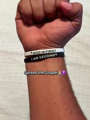 The image shows a close-up of a person's arm with a stack of bracelets. The arm is dark-skinned, and the hand is clenched into a fist. Three bracelets are visible. The top one is white, embossed with the words 'GOD IS FIRST' in black. The middle bracelet is black, with the words 'I AM SECOND' in white. The bottom bracelet is also black, devoid of any text. In the lower area of the image, a text overlay says 'Spread the Gospel' followed by a praying hands emoji and a purple cross emoji
