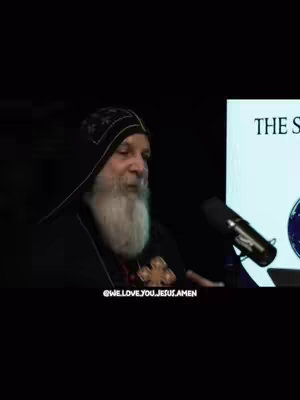 The image captures a man in a black robe, speaking into a microphone. He has a long white beard and is facing to the side. In the background is a screen with text. The overall setting is low-lit.