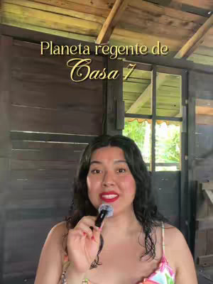 A woman with long, dark, wavy hair is holding a microphone. She is standing in front of a rustic wooden structure with sunlight filtering through. Text overlayed on the image at the top reads "Planeta regente de Casa 7" in a flowing, golden font.