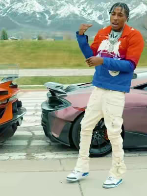 The image shows a man in a colorful jacket standing on a sidewalk with two luxury cars in the background, and mountains in the distance. The man is posing near the cars. He is wearing a red and blue jacket, white pants, and shoes. The scene conveys a modern and luxurious aesthetic.