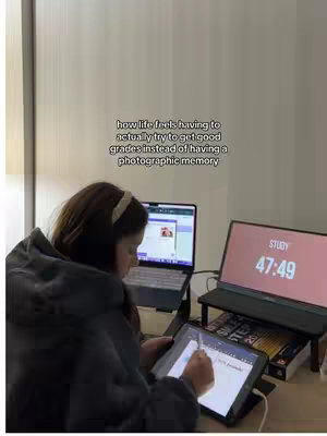 A woman is sitting at a desk with two laptops and a tablet in front of her. She is looking at the tablet and appears to be writing on it with a stylus. One laptop has a timer with the word "STUDY" and a countdown. The image has a muted color palette with soft lighting.