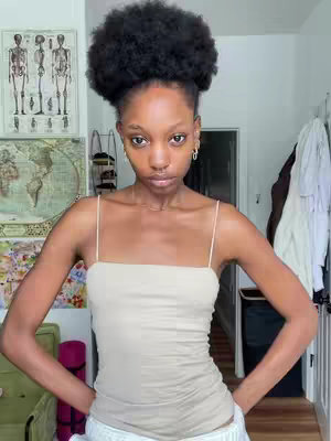 The image is a medium shot, featuring a woman with dark, tightly styled hair. She is centered and looking directly at the camera with a neutral expression. She is wearing a light-colored, spaghetti-strap top that emphasizes her bare arms. The background consists of walls and doorways within a room, suggesting a casual setting – the composition focuses on the woman’s upper body, creating a close-up effect.