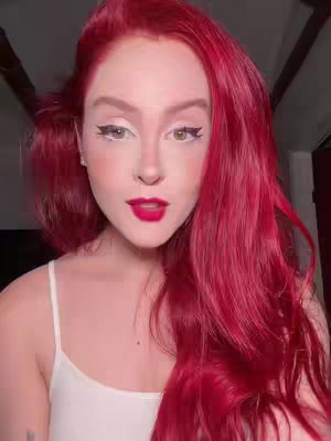 The image is a close-up of a woman with bright red hair and makeup. Her skin is pale, and she has a defined eyebrow arch, black eyeliner, and white eyeliner. Her lips are painted a deep red. She is wearing a white tank top. The background is dark and out of focus.