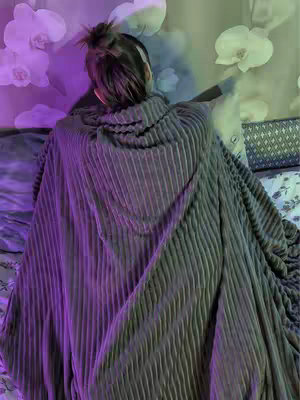 The image shows a person from behind, wrapped in a thick, ribbed blanket or shawl that drapes down. The background features blurred floral elements and a gradient of colors, with purple tones on the left and lighter hues on the right, suggesting a softly lit, intimate setting.