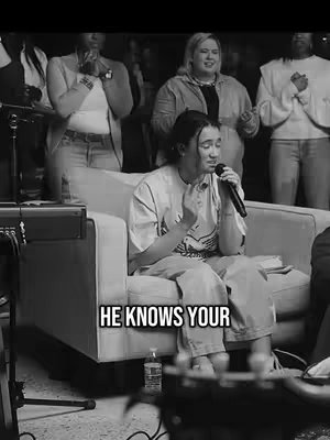 A black and white image featuring a woman with dark hair seated on a cream-colored armchair holding a microphone. She is dressed in a t-shirt and pants. Behind her are other people who appear to be in the background of a worship setting. Overlayed text states 'HE KNOWS YOUR'.