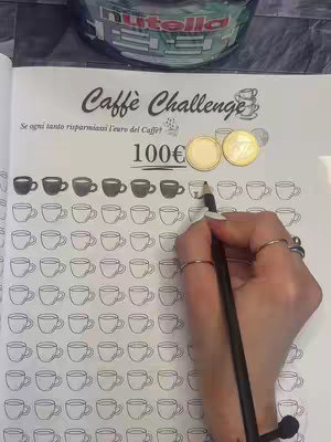 A person's hand is shown filling in a coffee cup on a printed page titled "Caffè Challenge." Two gold coins sit next to the cup chart, and there is a jar lid in the background.