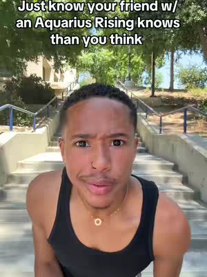 A man with short, dark hair stands on a set of outdoor stairs, facing the camera. He wears a black tank top and a gold necklace. The background includes trees, a building, and a bright sky, with text overlayed at the top.