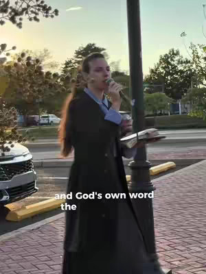 A woman standing in front of a street background. She holds a microphone in one hand and a bible in the other, with text overlaid on the bottom. The background includes a street, cars, trees, and a pole.