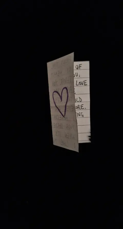 An image with a plain black background, with a piece of paper folded and held at an angle in the center. The paper is white with handwriting on it, and a purple heart is drawn in the middle of the paper. The handwriting is black and appears to be a personal message.
