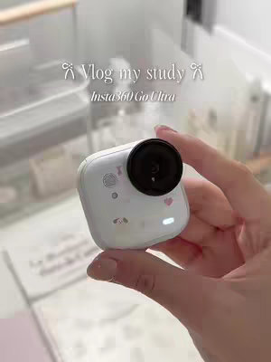 A close-up shot of a hand holding a white action camera, with a black lens. The camera has stickers with cute designs and a small light on. The background is a blurred, well-lit area, possibly indoors.