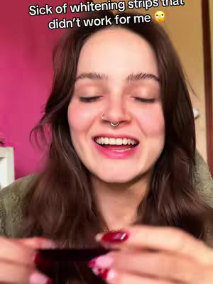 A close-up shot of a woman with long brown hair, wearing a green top, smiling with eyes closed, holding something that cannot be clearly identified in her hands. The background shows a pink wall and a part of a wooden frame.