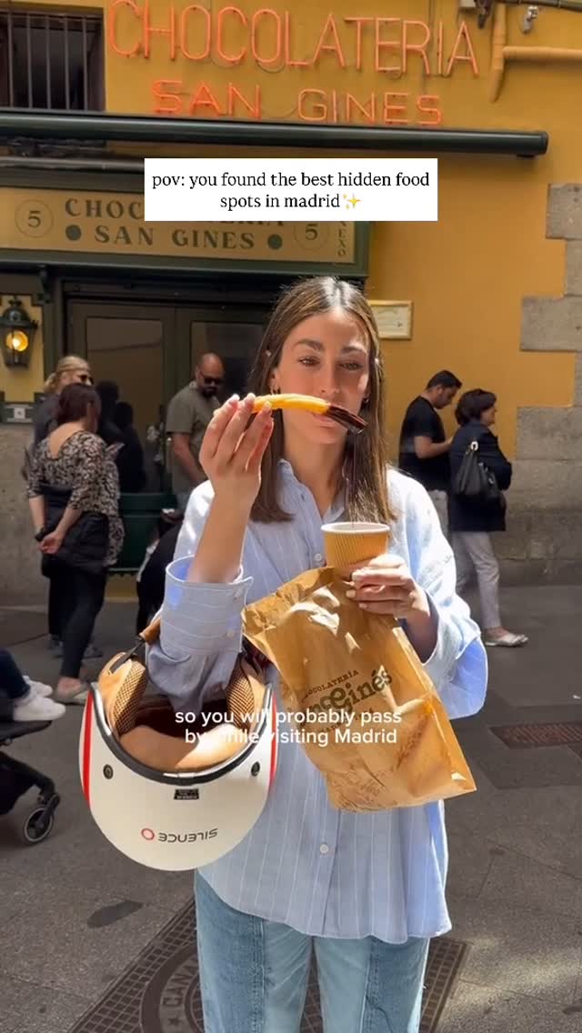 A woman stands in front of a building with a yellow facade and neon signage that reads 'Chocolateria San Gines', holding a churro dipped in chocolate in one hand and a to-go cup in the other. She has a helmet hanging by her side and paper bag from the shop. Other people are walking around in the background.