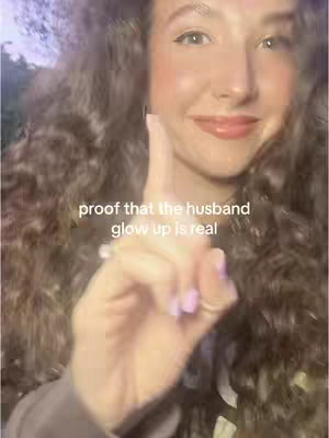 A woman with long, wavy brown hair is pointing her index finger up towards the right side of the frame. She is smiling with lipstick on. The background is blurred, but appears to be outdoors with some greenery visible.