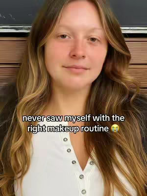A vertical shot featuring a woman with long, wavy, light-brown hair. She is wearing a white, button-up shirt. The background is a mix of indoor and outdoor elements, with a wooden panel and dark background above her. Text overlaying the image reads, 'never saw myself with the right makeup routine'.