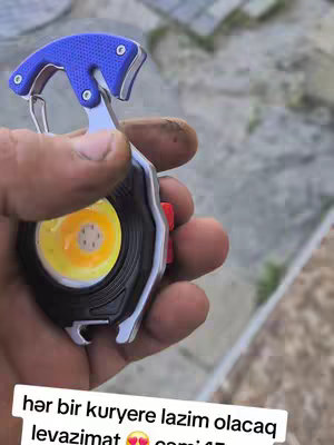 A close-up shot of a hand holding a multi-tool device, set against a blurred background. The device features a blue and silver handle, a black circular component with a bright yellow center, a red button, and a silver carabiner clip. The hand appears slightly dirty, suggesting practical use.