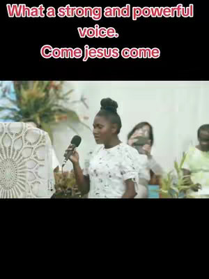 A vertical video still features a woman holding a microphone and singing in front of a crowd. The overlay text reads, 'What a strong and powerful voice. Come jesus come.' The woman is wearing a white top with floral patterns and her hair is styled in a bun. Behind her, people are visible, including some holding phones as if recording the performance. Green plants are in the background, with a white decorated structure to the left.