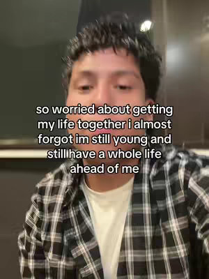 A selfie-style image featuring a young man in a checkered shirt against a blurred background. Text overlayed on the image reads, 'so worried about getting my life together i almost forgot im still young and still have a whole life ahead of me.' The composition is a medium shot focusing primarily on the person's face and upper body, with soft lighting.