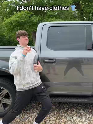 A young man stands next to a gray pickup truck, leaning against it casually. He's wearing a tie-dye hoodie, black pants, and white sneakers. The image has text overlay at the top