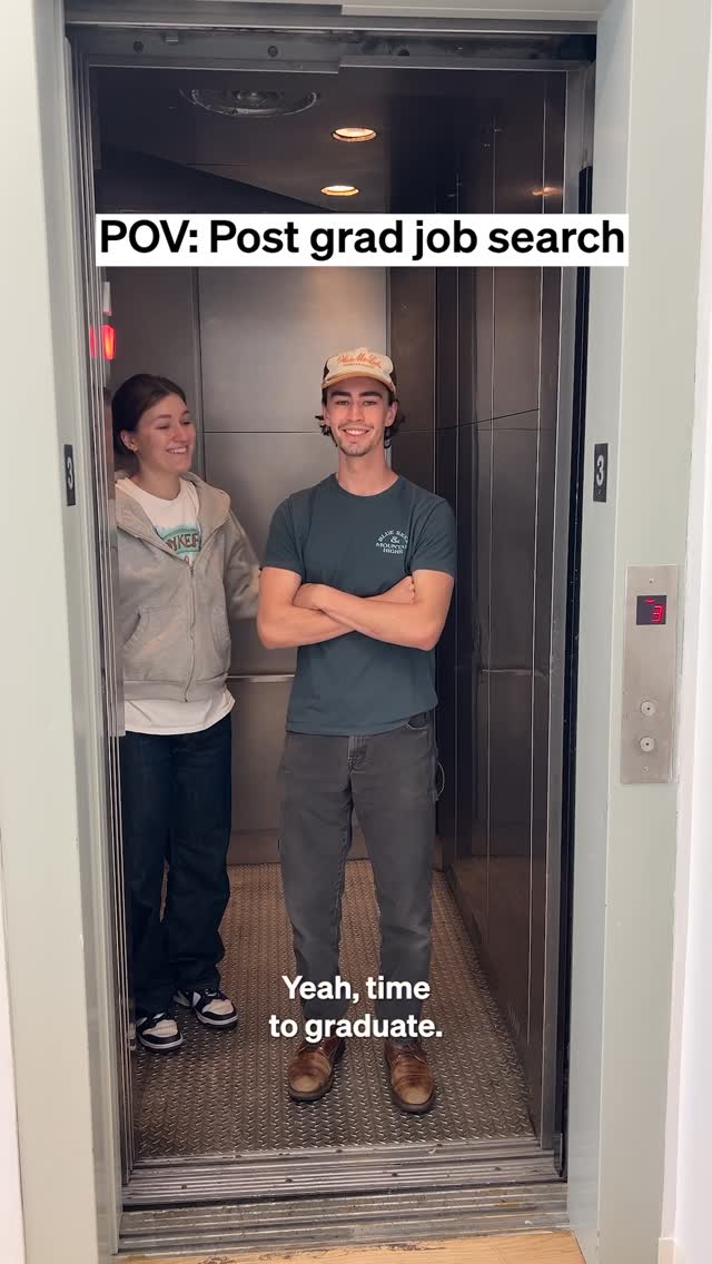 A man and a woman are standing in an elevator. The man is wearing a hat and a t-shirt, and the woman is wearing a hoodie. The elevator door is open and they are smiling.