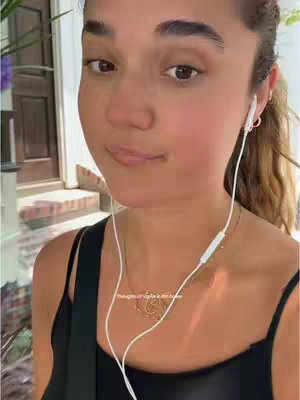 A woman wearing a black top and white earphones, is outside. She has light skin, brown eyes, and a necklace. The background includes a building entrance.