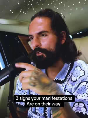 A man with a dark beard and long hair, wearing a blue patterned shirt, is positioned in front of a microphone. Text at the bottom of the image reads '3 signs your manifestations are on their way'. The background suggests an interior setting, possibly a car or studio, with a starry ceiling.