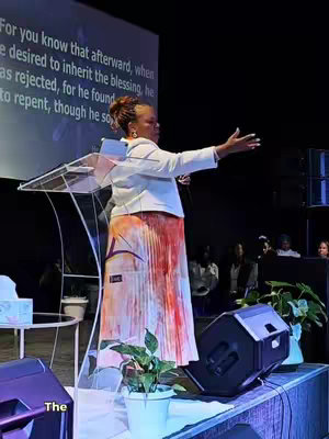 The cover image features a woman on a stage, likely a preacher or speaker, with a large screen behind her displaying text. She's wearing a white blazer over a colorful patterned skirt and is gesturing with one arm. The stage has a podium and small table with plants.