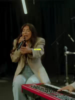 A woman singing into a microphone while sitting next to a red keyboard. She is wearing a beige blazer over light-colored pants. The background is dark with spotlights shining down.