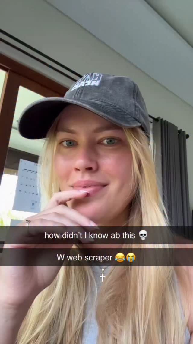 A selfie-style shot featuring a fair-skinned woman with long, blonde hair, wearing a gray baseball cap that partially obscures her forehead. She has blue eyes and is gazing at the camera with a slightly inquisitive look, her index finger touching her lower lip. The background is blurred, but hints at an indoor setting with wood paneling and drapes. Two lines of overlaid text appear towards the bottom of the frame with emojis.