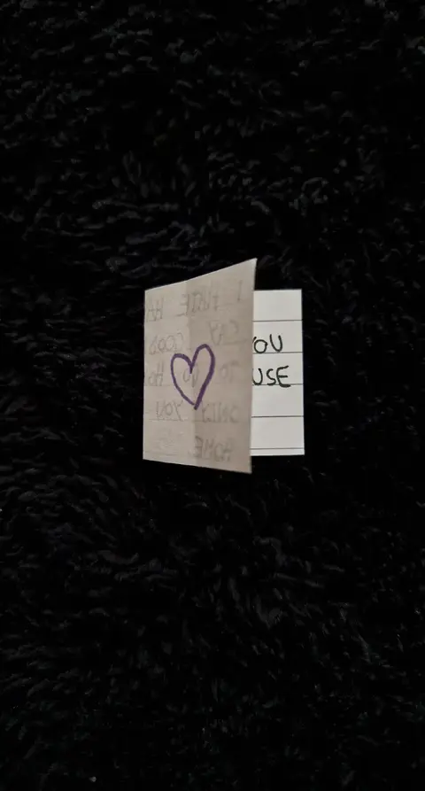The image shows a white note that is folded in half. A purple heart is drawn on the front half, with words written in the bottom left. The note is set against a black, fuzzy background.