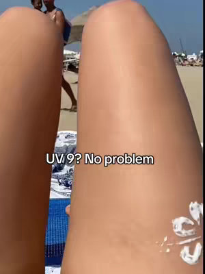 A close-up shot focuses on two tanned legs on a beach. The lower third of the legs are captured, featuring the knees and part of the thighs. Sunscreen is applied on the right leg in the form of flower. The background reveals a beach scene with other individuals in the distance. Text overlay reads "UV 9? No problem". The overall scene is bright and sunlit, evoking a summer vibe.