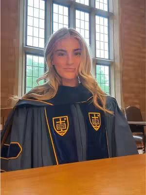 A woman in a graduation gown stands in a room with a large window behind her. The gown is dark blue with yellow accents and two patches with a crest. The woman has long blonde hair and is looking at the camera.