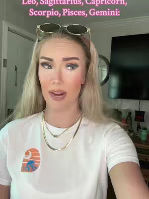 A medium shot shows a blonde woman with her hair over her shoulders. She wears sunglasses on her head, and is facing the camera, with her mouth open. In the background is a door and a television.
