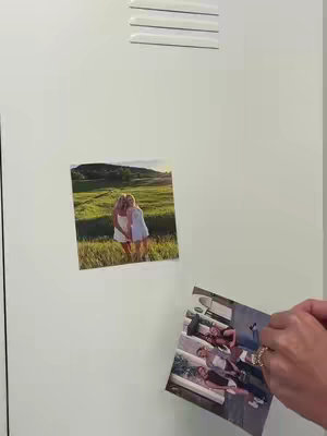 A light green locker door with three small vent slats at the top. Two photographs are attached to the door. The top photo shows two women embracing in front of a field with trees. The photo below is a group photograph, and one hand with rings is holding the photograph.