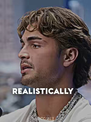 A medium shot of a young man in profile. He has light brown hair with blond highlights, styled with some volume. He has a goatee and stubble. The background is blurred but seems to be a professional setting. 'REALISTICALLY' is written in large, white text at the bottom.