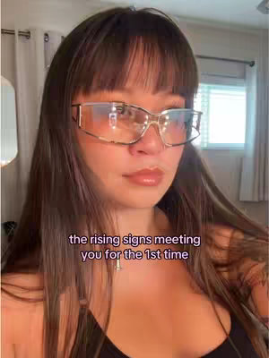 A selfie shows a woman with long brown hair, wearing large, rimless eyeglasses, and a black tank top. The background is slightly blurred with what appears to be curtains or drapes.