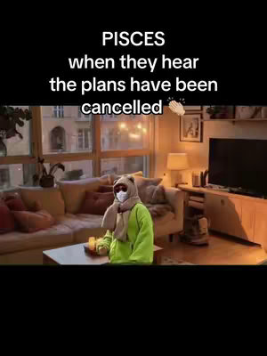 The image displays a cozy living room scene with a person in a green hoodie, mask, and scarf seated on a coffee table. The text overlay reads, 'PISCES when they hear the plans have been cancelled' above, suggesting a humorous take on a zodiac sign's reaction to changes in plans. A small clapping hands emoji is included. The lighting is warm, and the decor creates a comfortable, relatable setting.