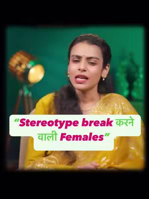 A woman with dark hair is centered in front of a blurry green background and a lamp. She wears a yellow patterned top and is speaking with a slightly open mouth. Text with a white background and black border spans across the image at the bottom. The text is in both English and an Indic script.