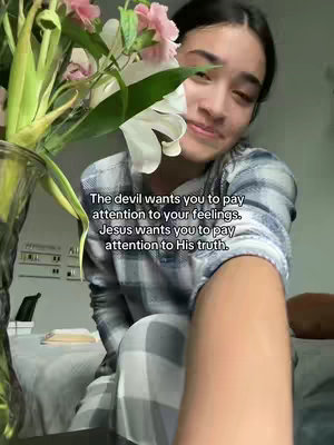 A selfie-style shot with a young woman in a plaid shirt partially visible, with a close-up focus. A vase with flowers is to her left, taking up about a third of the frame. The background is blurred, suggesting an indoor setting, with text overlaid on top.