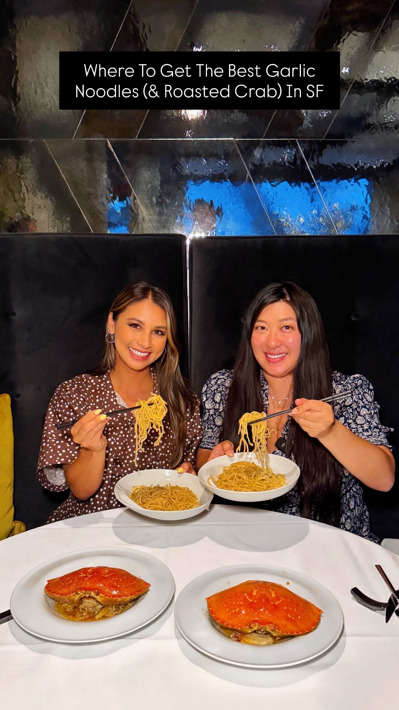 Two women are seated at a table, each holding a plate of garlic noodles and smiling. In front of them are plates with roasted Dungeness crab. The background features a dark, textured wall. A black rectangular banner at the top of the image displays white text.