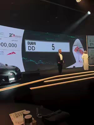 The image shows a large stage with a dark-colored background. A man in a tuxedo stands at center stage, facing away from the camera. Behind him is a large screen displaying varying information, including license plate information. The stage is lit with spotlighting, and there are other individuals visible on the stage, including a presenter. There are podiums in the foreground, with some items on them. The overall composition suggests a formal, public event.