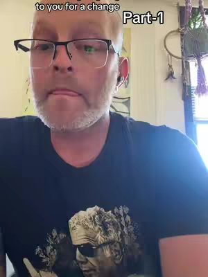 A middle-aged man with glasses is the focal point, his face close and centered. His expression is serious, with his tongue touching his lips. He's wearing a black shirt with an image, and the background has some visible indoor elements. The lighting seems natural.