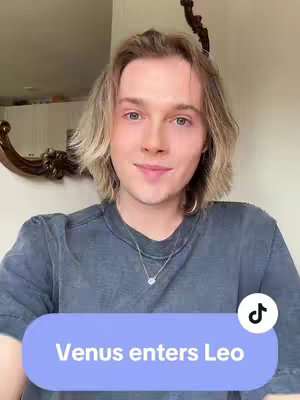 A person with fair skin and blonde hair smiling at the camera. The text overlay declares \"Venus enters Leo\". In the background visible is a mirror and a part of the wall.