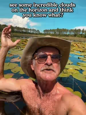 A man with a hat and sunglasses stands in water filled with lily pads. The sky is a brilliant blue with a few clouds in the background.