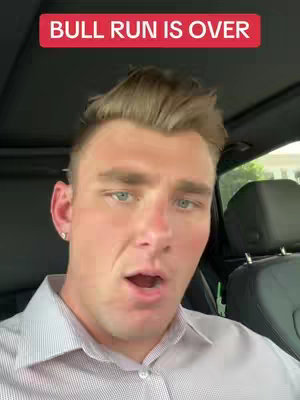 A man with light skin sits in the driver's seat of a car, facing the camera. He has short, light brown hair and is wearing a light colored shirt. A red banner with white text, 'BULL RUN IS OVER', is placed above his head. The background shows the car's interior.