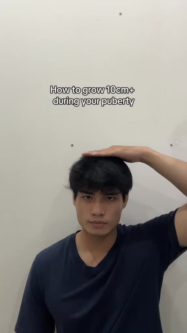 A young man stands in front of a plain, off-white wall. He is wearing a dark blue t-shirt and has dark hair. The caption on the image reads, "How to grow 10cm+ during your puberty." He is holding his right hand flat above his head, as if measuring the height of his hair.