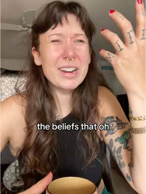 A woman with long brown hair is in a close-up shot, looking off to the side with an expressive, somewhat distressed face. She has a nose ring and a few tattoos on her arms. The background appears to be a domestic setting.