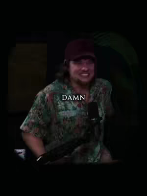 A man with long hair and a maroon cap is captured mid-sentence. The word 'DAMN' is overlaid on the image in bold white text. The man is wearing a green shirt with a floral print. The background is dark and out of focus.