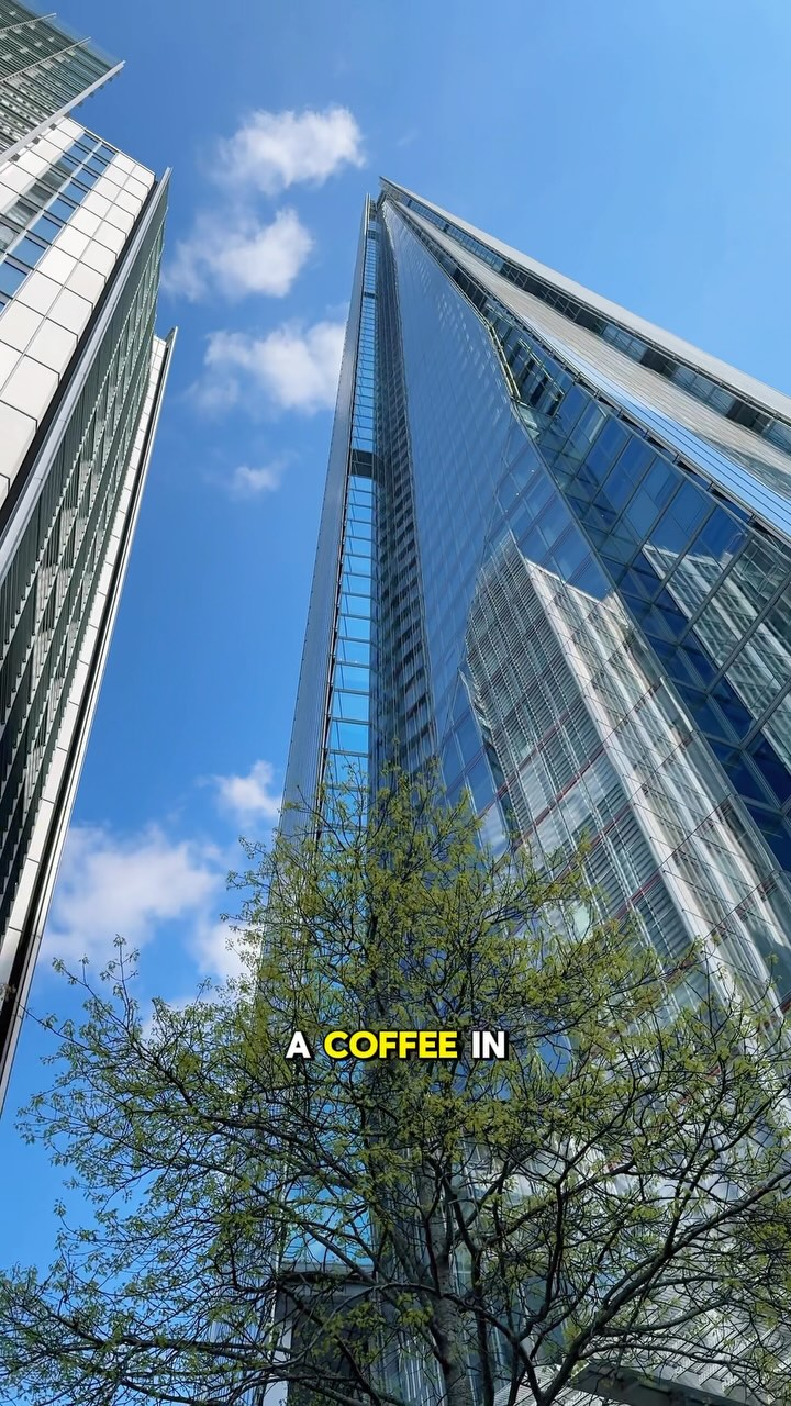Looking up at the Shard skyscraper in London from ground level on a clear, sunny day. The image is taken from a low angle, emphasizing the height and grandeur of the building. The sky is a vibrant blue with few scattered clouds. In the foreground, green tree branches and leaves frame the bottom of the image.