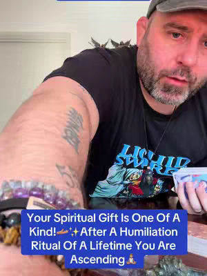 A close-up shot focuses on a bearded man with a gray cap, displaying a text overlay in a blue rectangular box at the bottom. The text reads, 'Your Spiritual Gift Is One Of A Kind!', followed by the sentence, 'After A Humiliation Ritual Of A Lifetime You Are Ascending'. The man's arm, adorned with tattoos, is in view, as are bracelets and a table strewn with tarot cards, and a bottle of a blue substance sits beside him.
