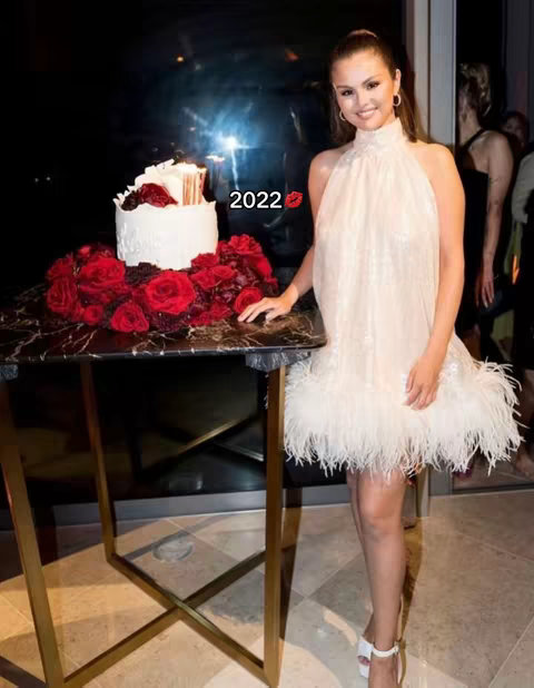 A woman in an off-white dress poses in front of a table with a cake and red roses. Behind her, there's a dark background and other people. The hook in the image has the numbers and a kiss emoji.